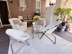 Un patio con una silla blanca acolchada, una tumbona, una mesita con un florero y un refrigerador.