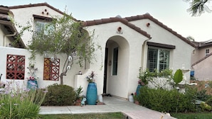 A house with a white exterior, brown roof, and a number '16164' on the front. There is a tree in the front yard, a cat walking on the porch, and various potted plants and shrubs around. - Your cozy getaway awaits in San Diego at this delightful house. (San Diego)