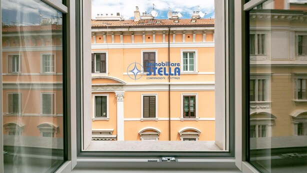 Ikkunasta avautuu näkymä parvekkeelliseen rakennukseen, taivas on kirkas ja näkyvissä on yrityksen »Immobiliare Stella» logo.