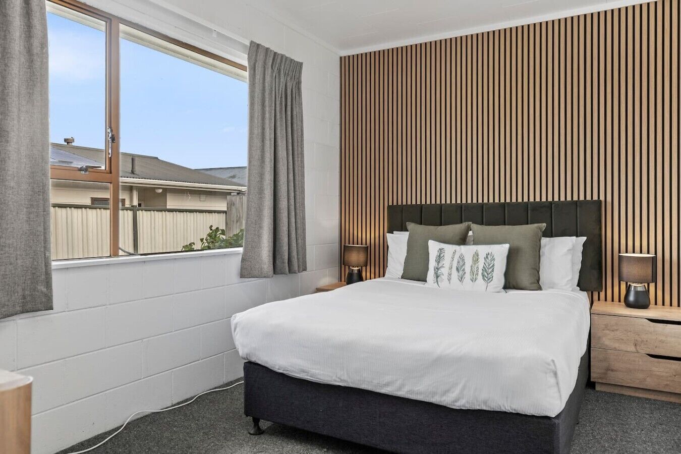 A bedroom with a bed, two bedside tables, a window with curtains, and a wooden slat wall.