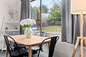 Uma área de refeições com mesa de madeira, cadeiras pretas, um vaso branco com flores e vista para o verde lá fora.