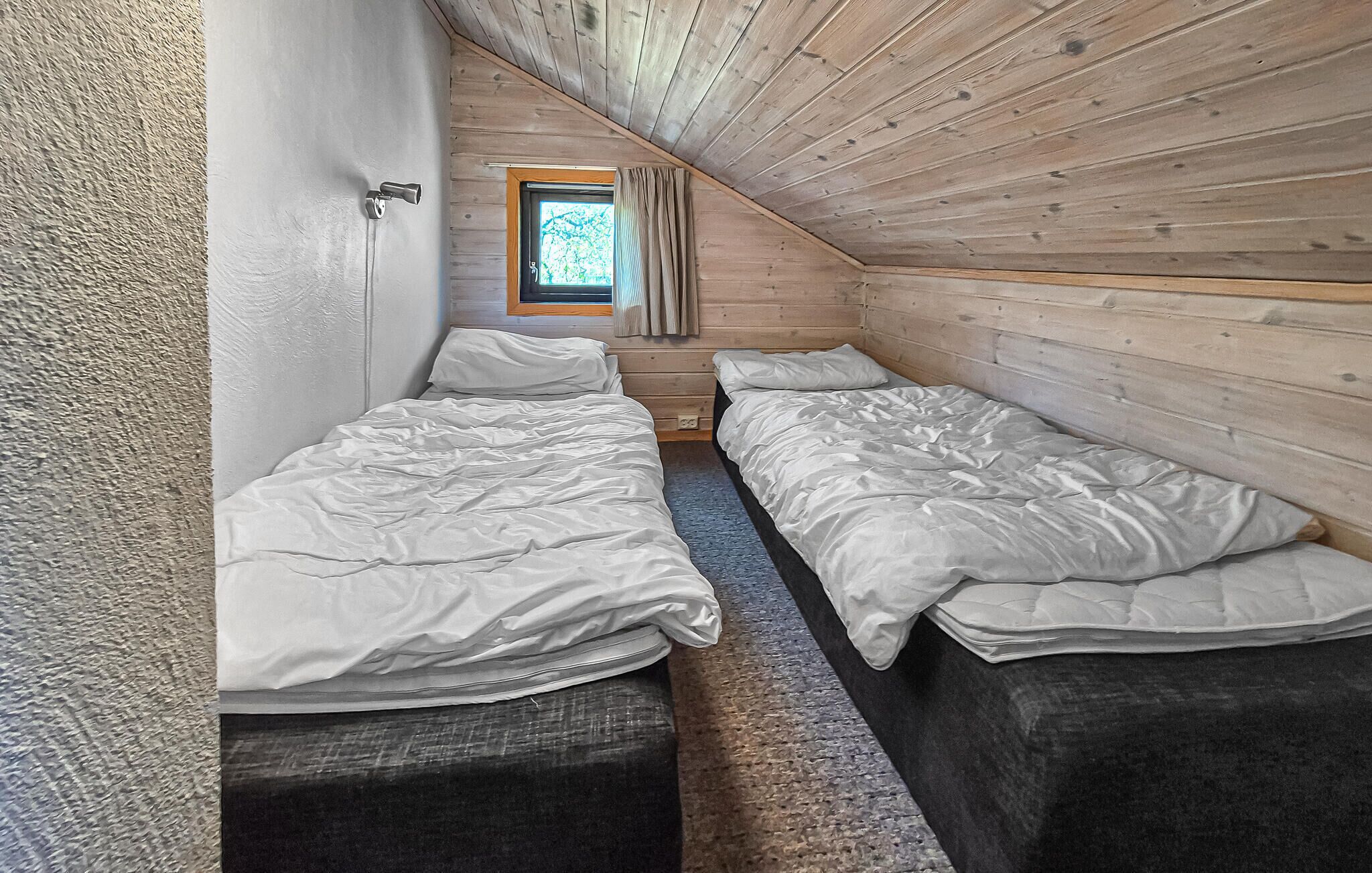 A small, cozy room with two single beds, a window with curtains, and a wooden ceiling.