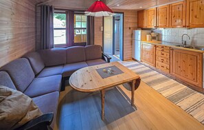 Ein gemütliches Blockhaus-Interieur mit einer Couch, einem kleinen Holztisch, einer Küchenzeile mit Holzschränken und einer roten Hängeleuchte.