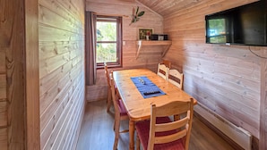 Un intérieur en bois comprenant une table à manger, des chaises, un téléviseur fixé au mur et une fenêtre donnant sur un paysage verdoyant.