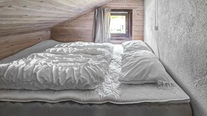 Ein Einzelbett in einem Raum mit Holzwänden und einer geneigten Decke, ein Fenster mit Vorhängen und eine strukturierte Wand auf der rechten Seite.