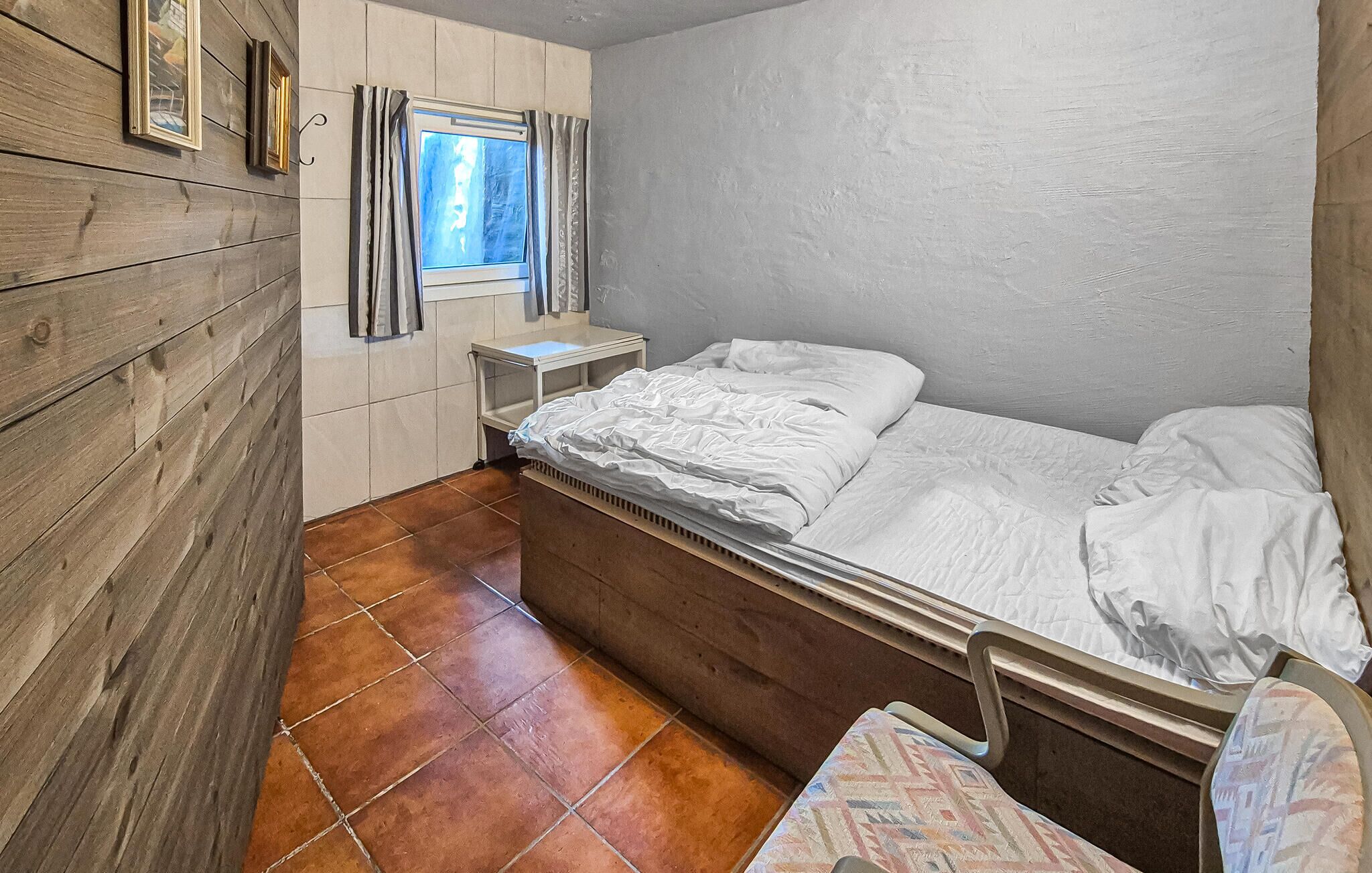 A small, compact room with a wooden wall, a single bed, a nightstand, a window with curtains, and a tiled floor.