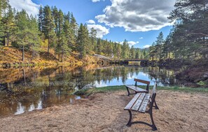 A bench by a calm river with trees and a bridge in the background.