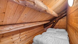 Interior de cabana de madeira com teto inclinado, cama com roupa de cama branca e uma tomada elétrica visível.