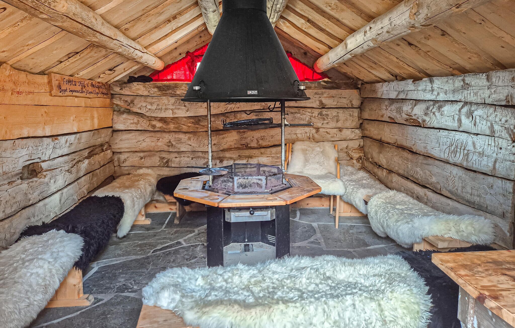 A log cabin interior with a central fireplace, stone flooring, and fur rugs.