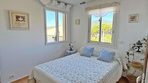 A bedroom with a bed, two windows, a small table with a vase, and a lamp on a side table. - Apartment T4 Duplex - 6 people - Sea view - Swimming pool residence - Air conditioning - WiFi -... (STE MAXIME)