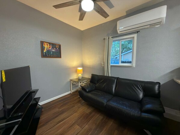 Un salon avec un canapé en cuir noir, un meuble pour la télévision, une lampe, un ventilateur de plafond et un appareil de climatisation.