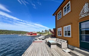 A wooden building with a dock extending into a body of water, a red house in the background, and a clear sky. - Amazing home in Mandal with house sea view (Mandal)