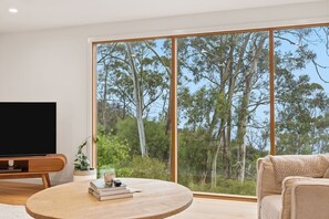 A modern living room with a large window, a flat-screen TV, a round wooden table, and a sofa. - Bush Escape with Water Views (Tinderbox)