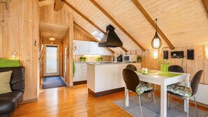 Ein gemütliches Blockhaus-Interieur mit Holzvertäfelung, einem Essbereich mit Tisch und Stühlen, einer Küche mit weißen Schränken sowie einem Schlafbereich im Zwischengeschoss mit geneigter Decke.