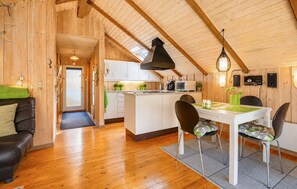 Ein gemütliches Blockhaus-Interieur mit Holzvertäfelung, einem Essbereich mit Tisch und Stühlen, einer Küche mit weißen Schränken sowie einem Schlafbereich im Zwischengeschoss mit geneigter Decke.