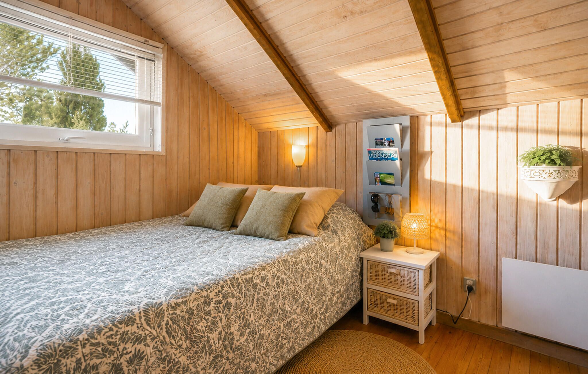 Ein Schlafzimmer mit einem Bett, einem Nachttisch, einem an der Wand befestigten Regal mit Büchern und einem Fenster mit Jalousien.