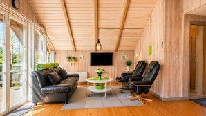 Ein gemütliches Zimmer mit Holzbalkendecke, einem Sofa, zwei Sesseln, einem Fernseher und einem kleinen Tisch mit Pflanzen.