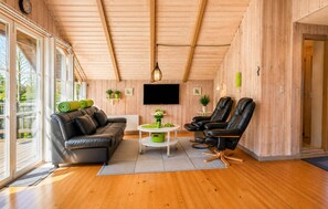 Ein gemütliches Zimmer mit Holzbalkendecke, einem Sofa, zwei Sesseln, einem Fernseher und einem kleinen Tisch mit Pflanzen.