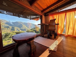 Un intérieur en bois avec une table, une chaise, un tapis et une vue sur les montagnes, visible à travers de grandes fenêtres.