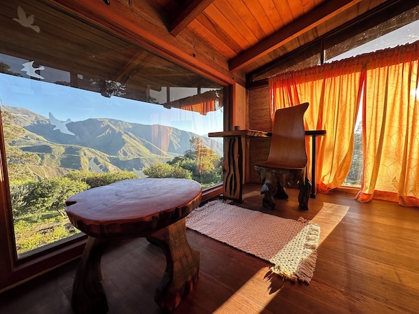A wooden interior with a table, a chair, a rug, and a view of mountains through large windows.