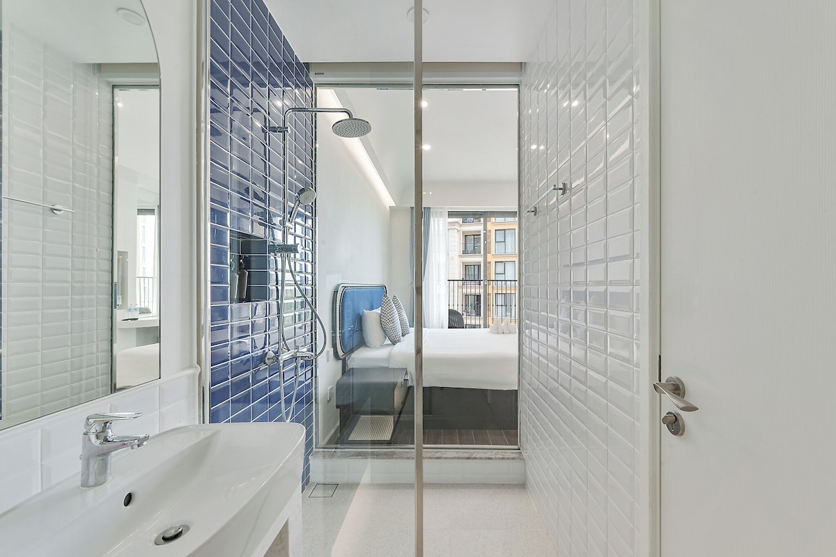 A modern bathroom with a glass shower enclosure, a white sink, and a visible bed in the adjacent room.