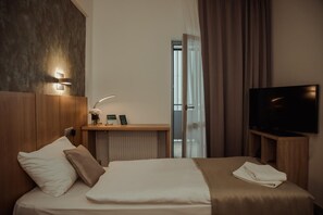 A hotel room with a bed, a television, a bedside table, a nightstand, a window with curtains, and a wall-mounted light fixture. - Hotel Arena (Kosice)