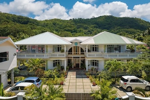 A two-story building with a central entrance, surrounded by a garden with palm trees, and a hilly landscape in the background. - 340 Degrees Mountain View Apartments (Mahé Island)