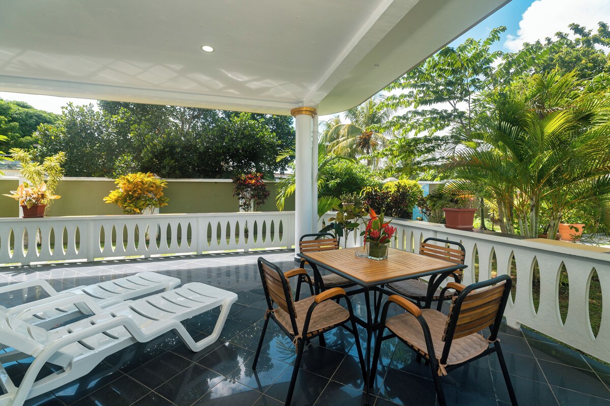A covered outdoor patio with a dining table and chairs, surrounded by potted plants and a white railing.