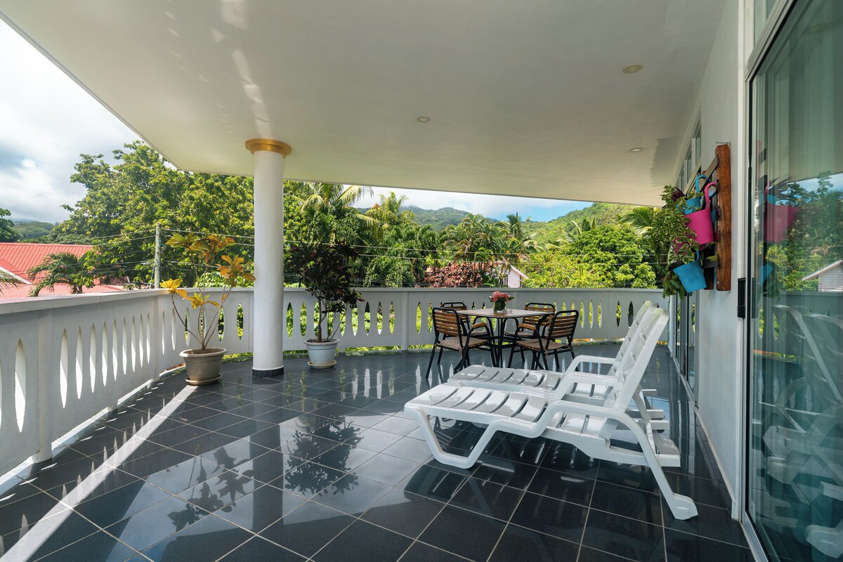 A balcony with a tiled floor, white lounge chairs, a table with chairs, potted plants, and a view of greenery and mountains.