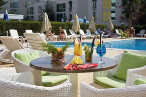 Zona junto a la piscina con sillas de mimbre, una mesa con bebidas y vistas a la piscina y al edificio al fondo.