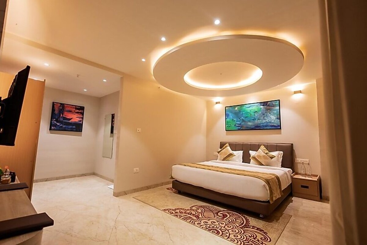 A modern hotel room with a large bed, a flat-screen TV, a circular ceiling light, and a painting on the wall.