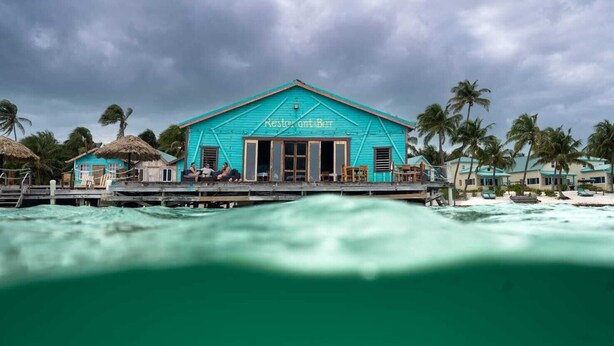 Un edificio turquesa con un cartel que dice 'Restroom & Bar' sobre un muelle con palmeras y una playa al fondo.
