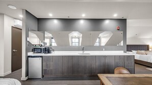 Una habitación de hotel moderna con kitchenette, un espejo grande, una cama con cabecera y un pequeño rincón de descanso.
