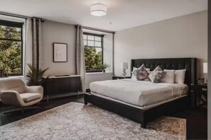 A bedroom with a large bed, a chair, a desk, and a window with a view of trees. - The British Hotel (Gatineau)