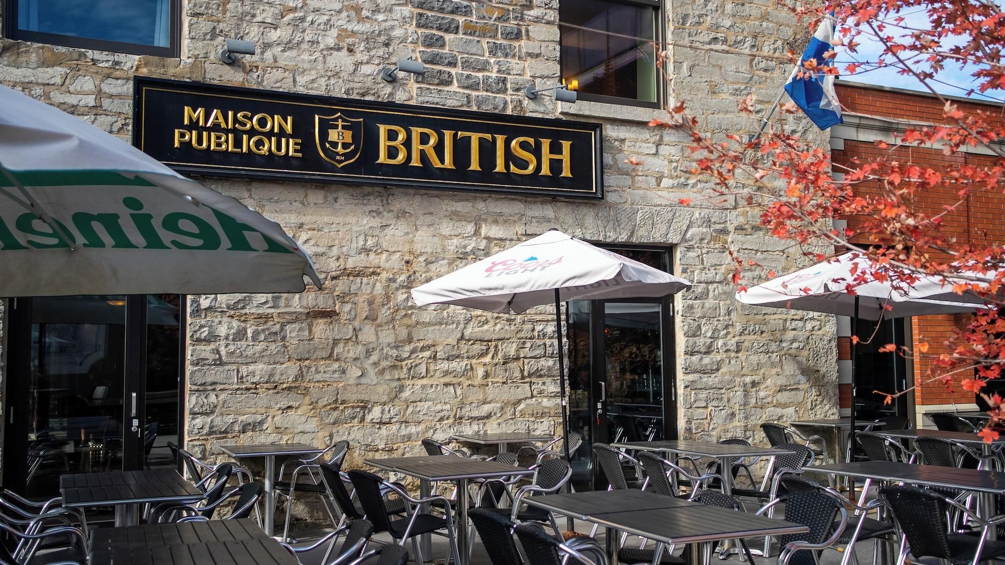 A stone building with outdoor seating, a sign reading 'MAISON PUBLIQUE BRITISH,' and a flag.
