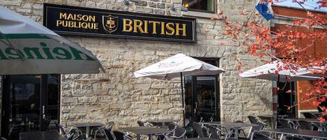 A stone building with outdoor seating, a sign reading 'MAISON PUBLIQUE BRITISH,' and a flag.
