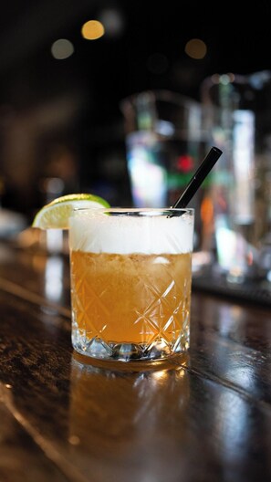 A glass of amber-colored beverage with a lime wedge and a straw, placed on a wooden surface. - The British Hotel (Gatineau)