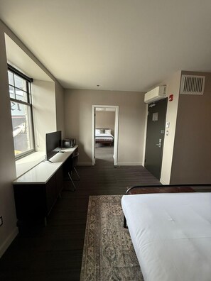 A hotel room with a bed, desk, chair, window, and a door leading to another room. - The British Hotel (Gatineau)