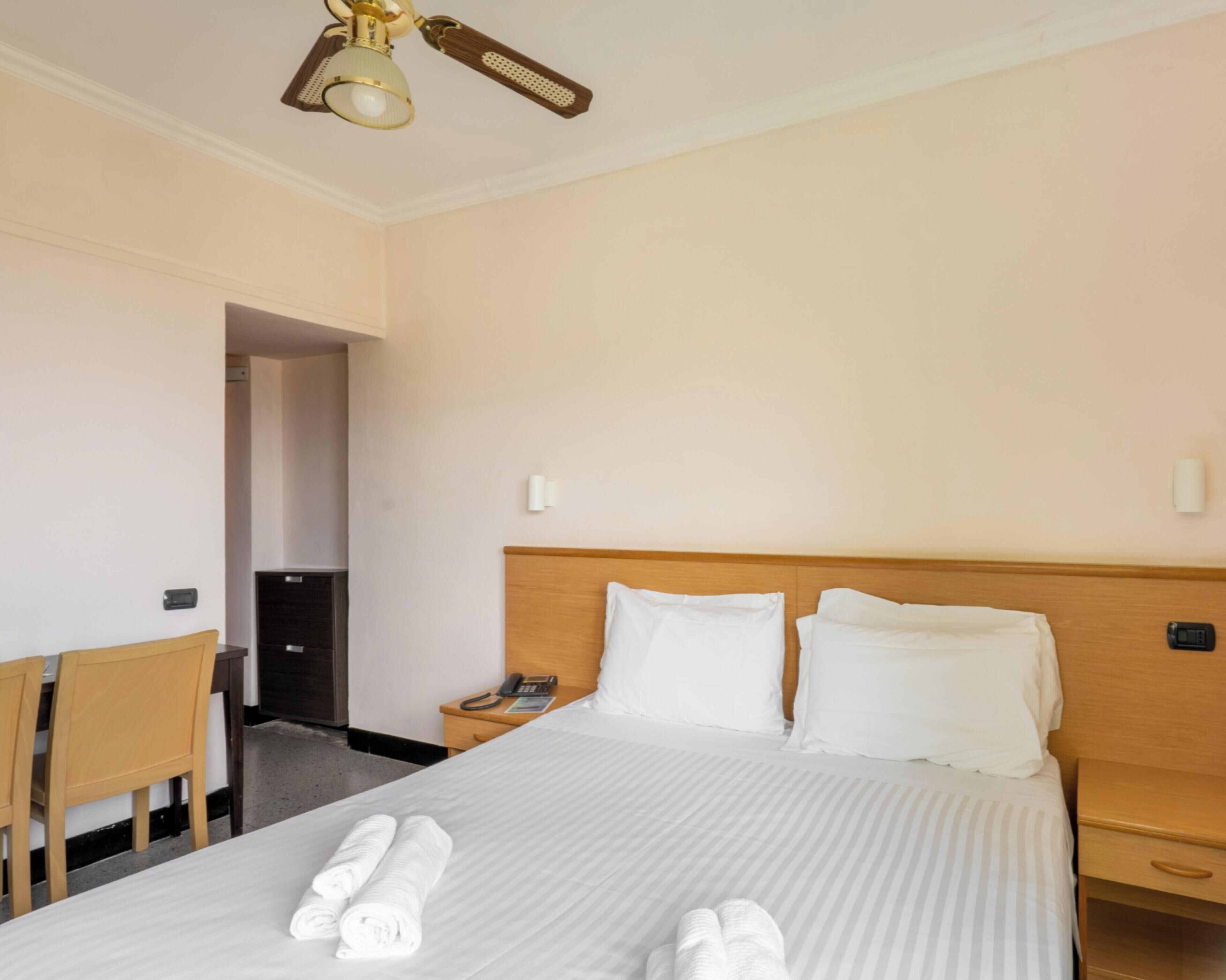 Une chambre d’hôtel équipée d’un lit, d’un bureau, d’une chaise, d’une table de chevet, d’un téléphone et d’un ventilateur de plafond.