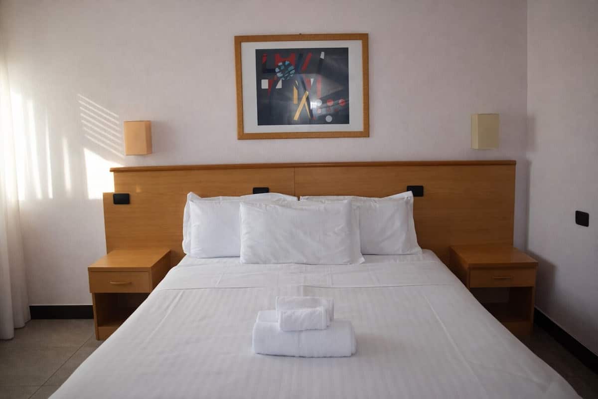 Habitación de hotel con una cama grande, dos mesitas de noche, un cuadro abstracto enmarco y una ventana con cortinas.