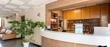 A reception desk with a wooden finish, a potted plant, a sofa, and a bookshelf with various items.