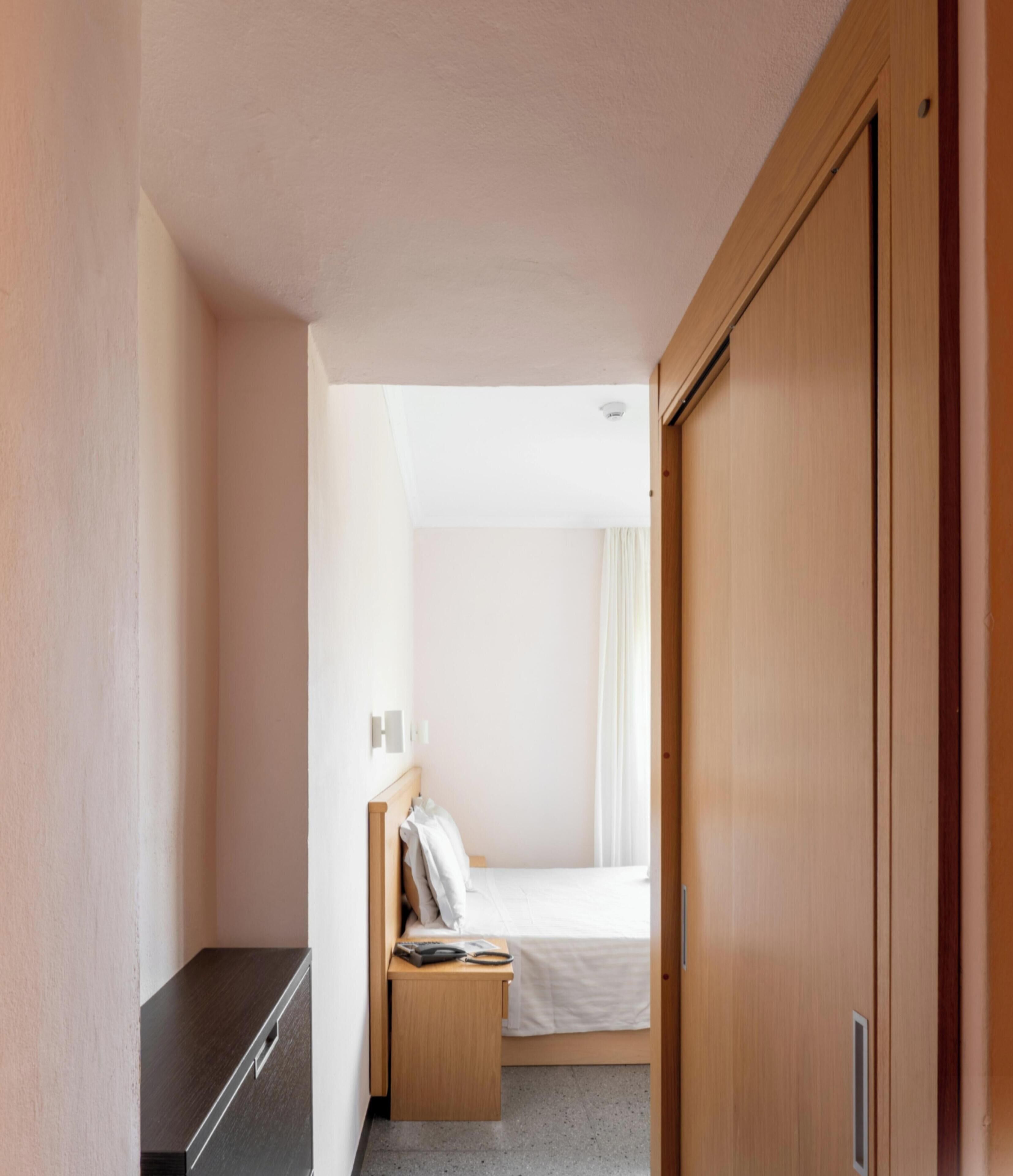 A narrow hotel room with a bed, bedside table, and a wardrobe.