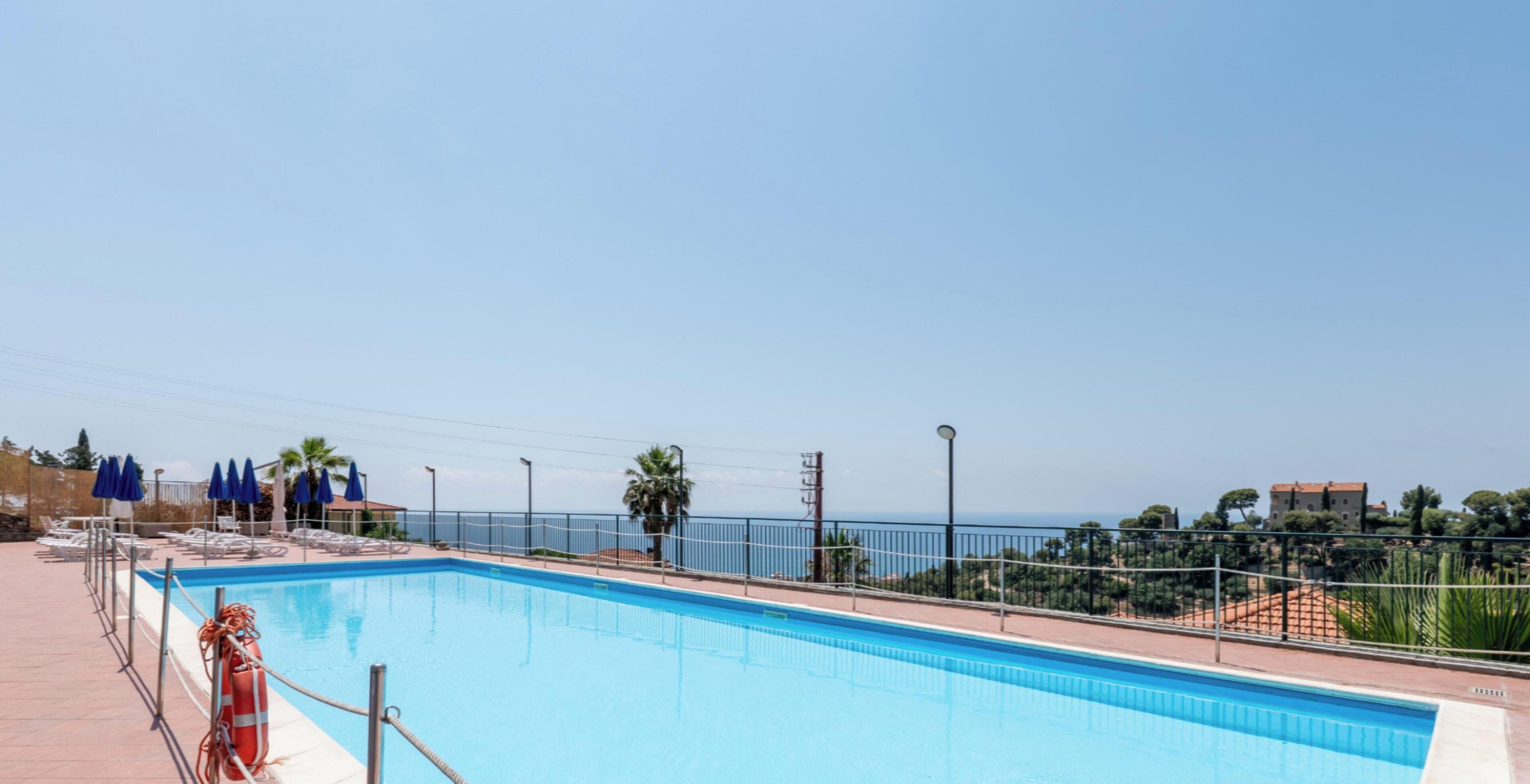 Una piscina con vistas claras al mar y un área vallada con sillas de descanso.
