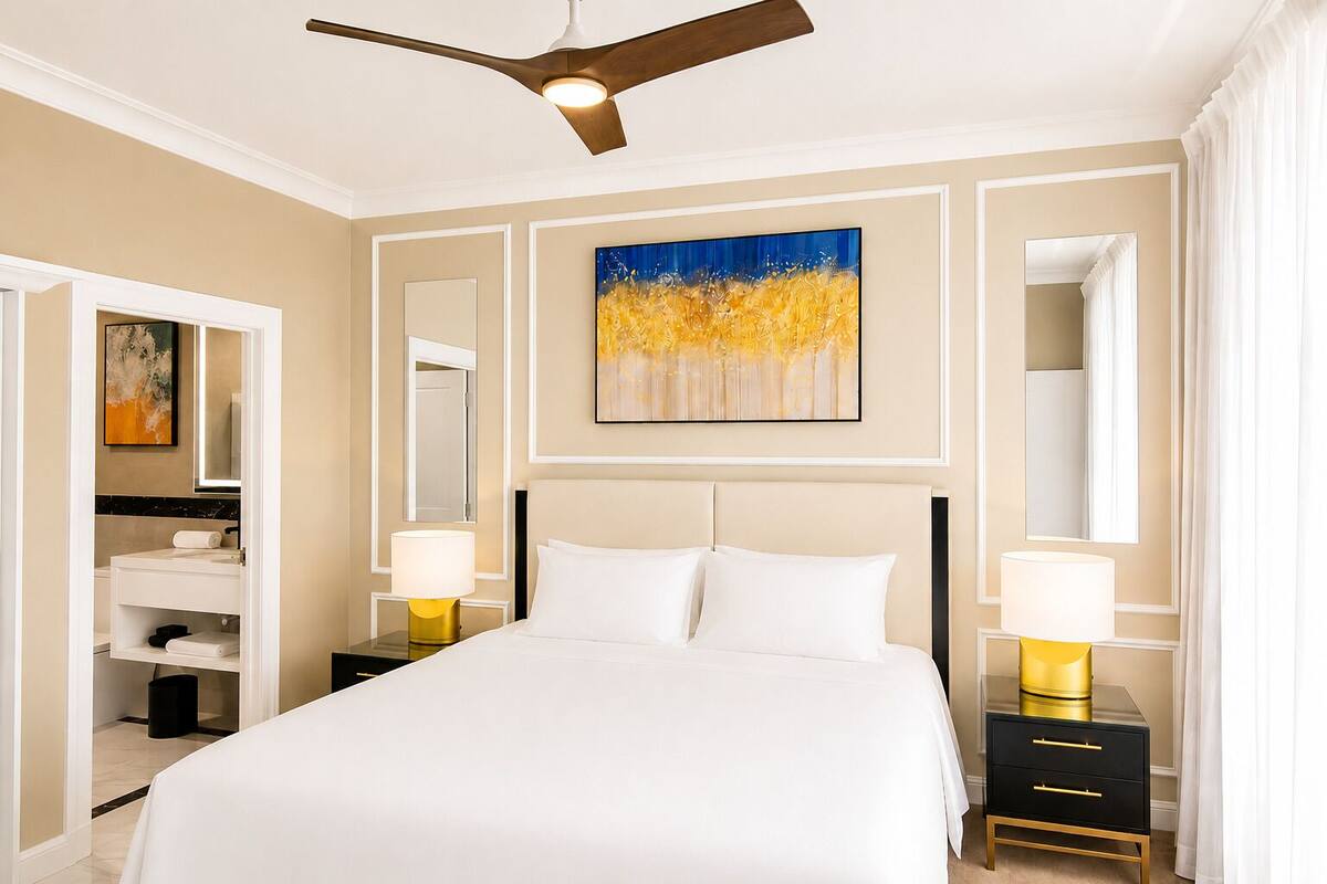 A hotel room with a bed, two bedside tables with lamps, a ceiling fan, a painting, and a bathroom with a mirror and sink.