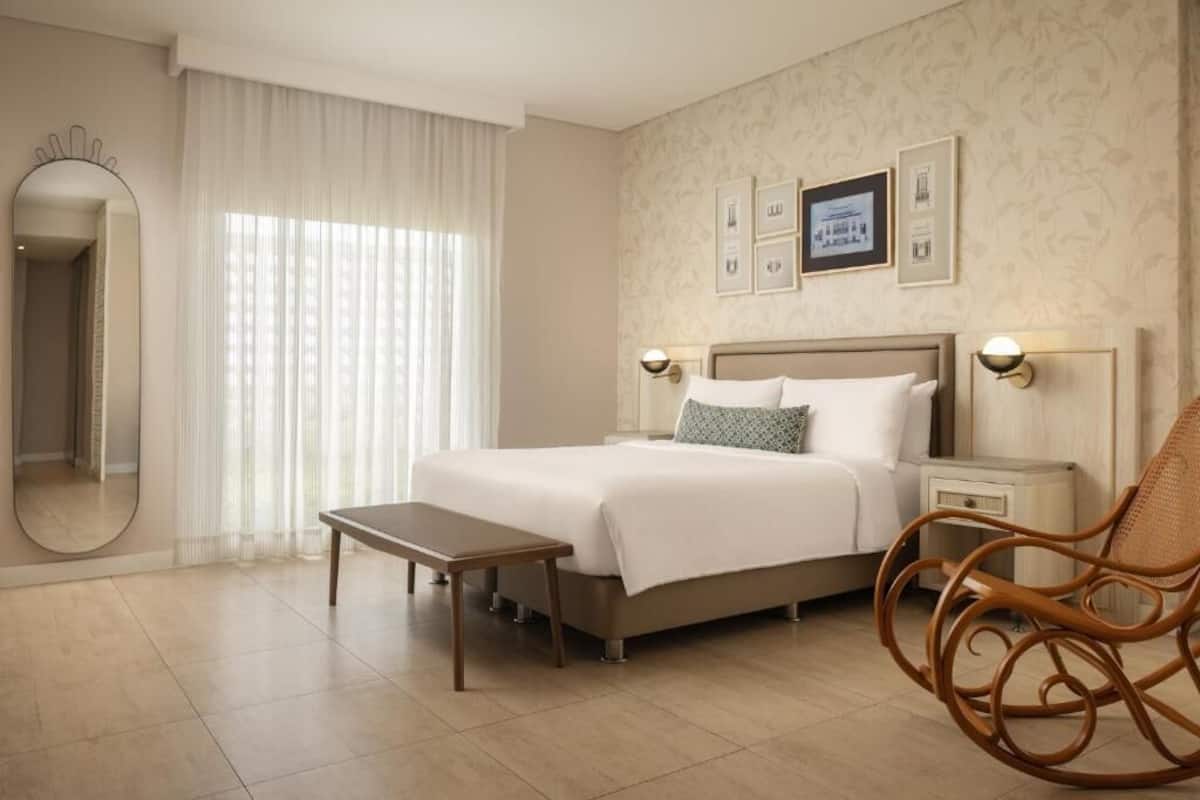 Una habitación de hotel con cama, mesitas de noche, una silla, un espejo y una ventana con cortinas.