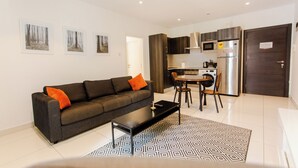 Een moderne woonkamer met een bank, salontafel en een eethoek met stoelen. Er is een keuken met een koelkast, magnetron en kastjes.