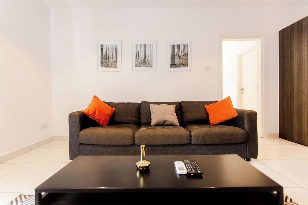 A modern living room with a grey sofa, orange cushions, a black coffee table, and three framed pictures on the wall.