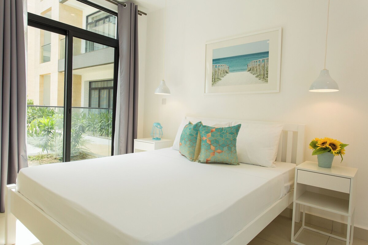 A bedroom with a large bed, a nightstand with a vase of sunflowers, a framed picture of a beach, and a view of a balcony with plants.