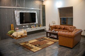 A living room with a sofa, a flat-screen TV, a cabinet, a lamp, and a window with blinds.