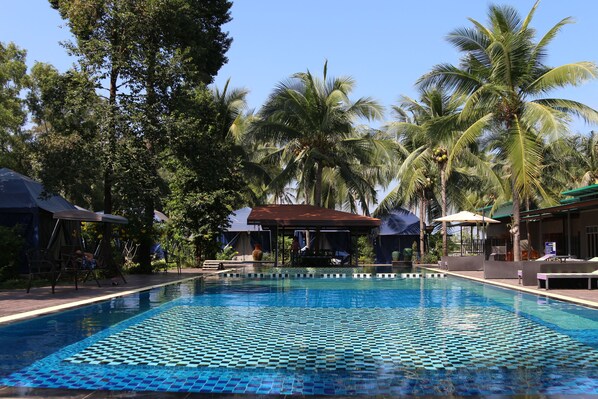 A swimming pool with tiled patterns, surrounded by palm trees and shaded areas.